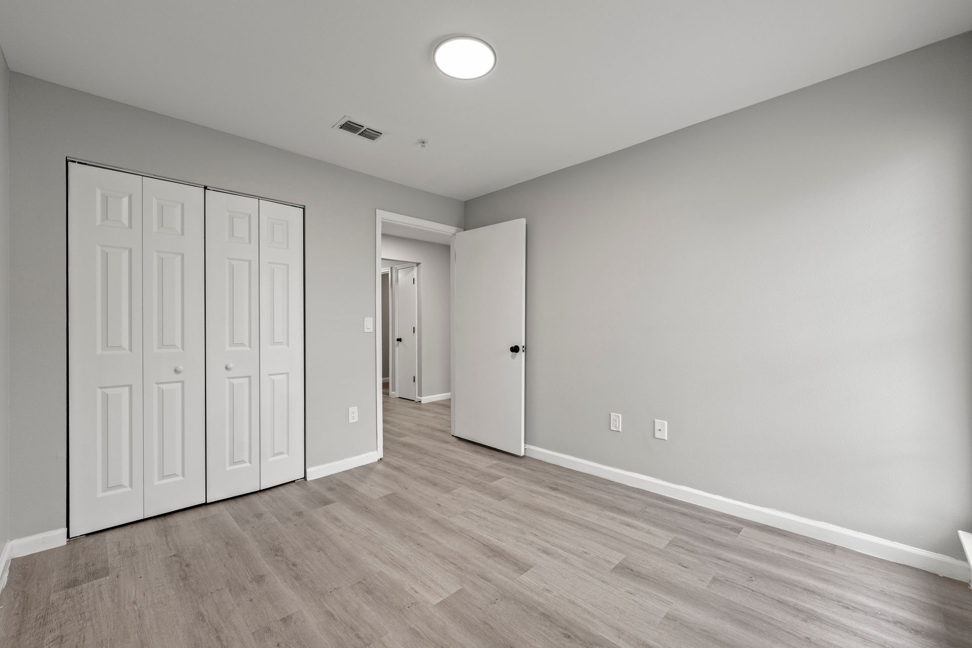 Empty gray bedroom with white closet doors, open door, and light wood flooring.