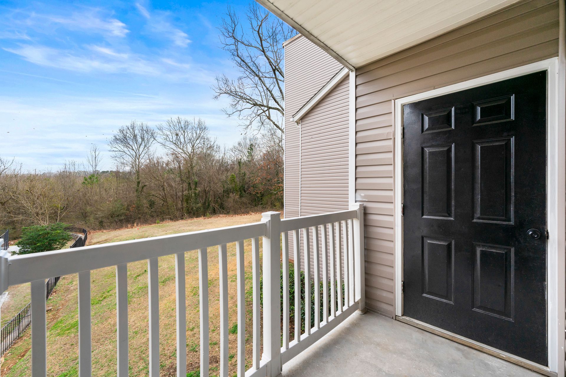 Front porch with white railing, black door, and a view of trees and grass beyond.