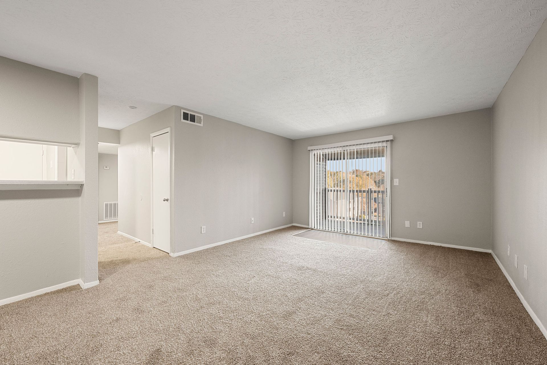 Empty beige carpeted living room with white walls and a sliding glass door to a balcony.