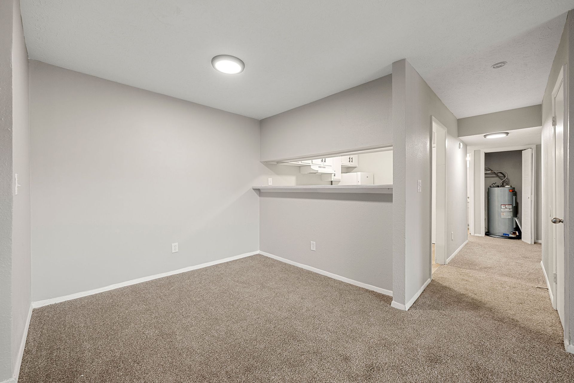 Empty carpeted hallway and room with white walls, gray ceiling light, and open pass-through to kitchen.