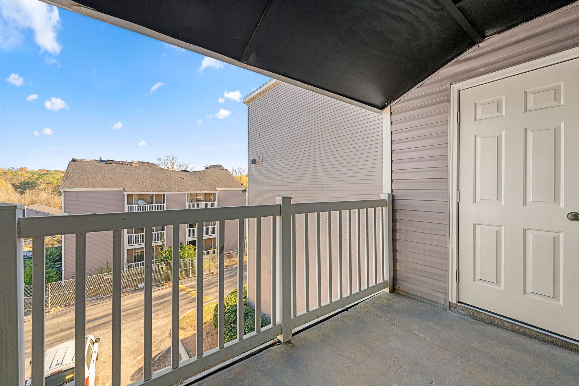 Balcony with gray railing, beige siding, white door, and a view of rooftops and trees.