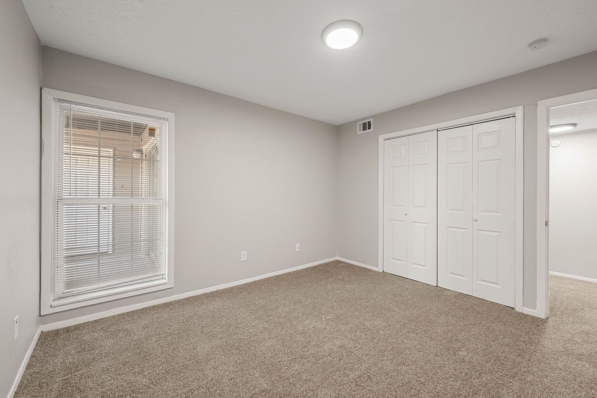 Empty carpeted bedroom with gray walls, a window, closet doors, and a ceiling light.