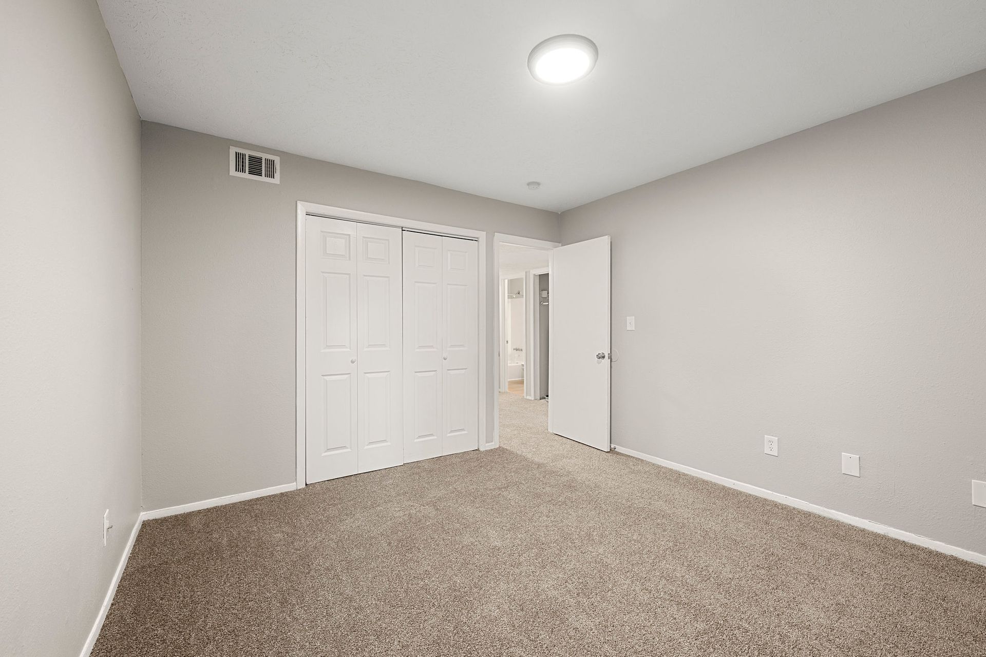Empty bedroom with gray walls, beige carpet, white closet doors, and an open doorway.