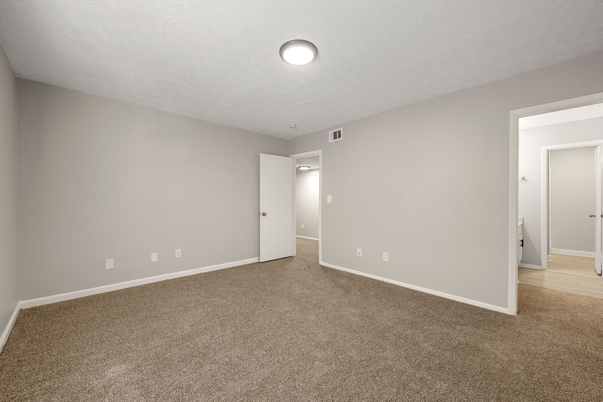 Empty beige carpeted room with white trim, ceiling light, and open doorway to a hallway and bathroom.