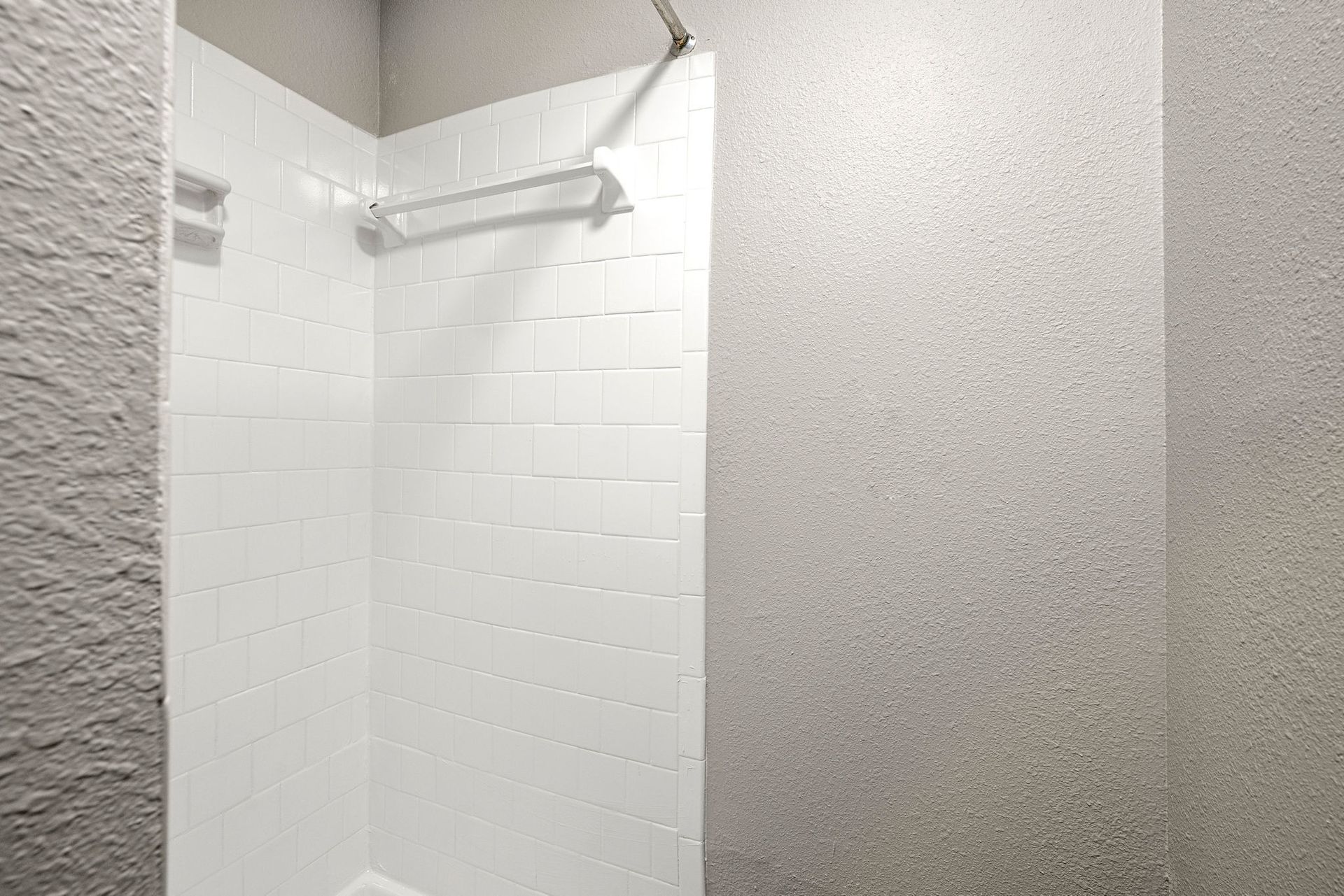 Small tiled shower stall with white walls and a curtain rod, beside a textured gray wall