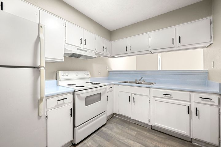 Bright white kitchen with stove, refrigerator, and L-shaped cabinets with blue backsplash.