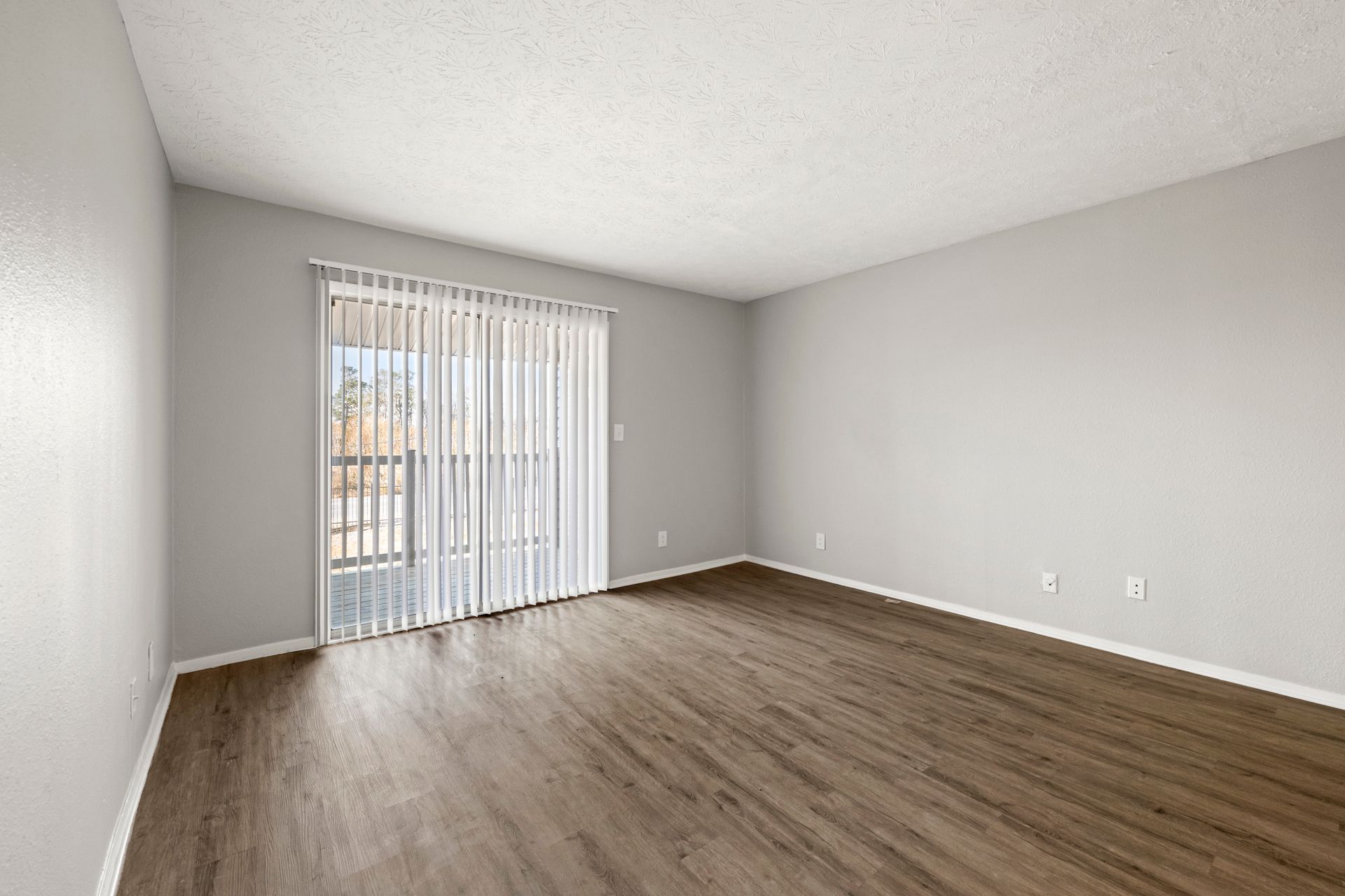 Empty gray living room with wood flooring and sliding glass door with vertical blinds