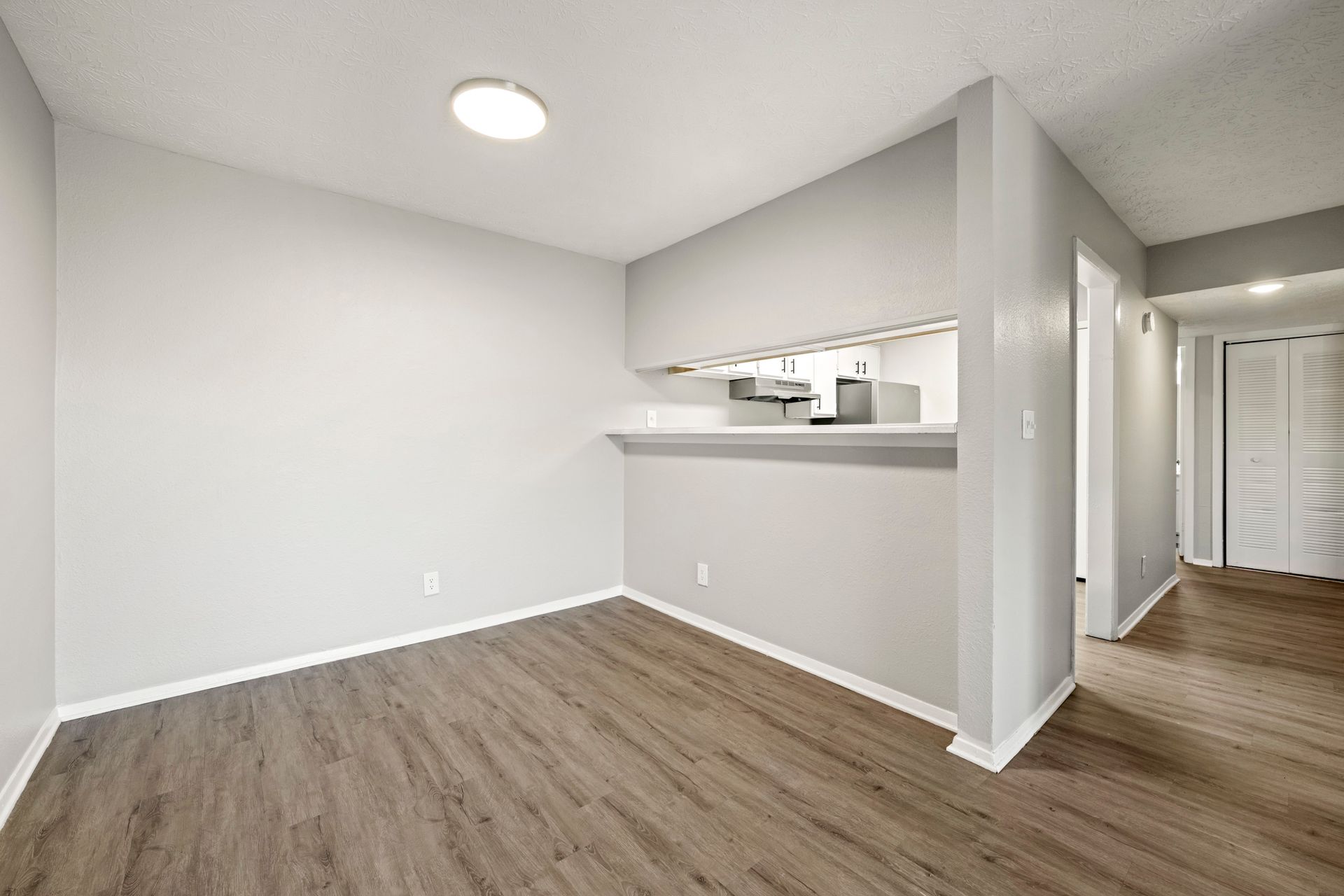 Empty white-and-gray apartment room with wood floors and a pass-through counter to the kitchen