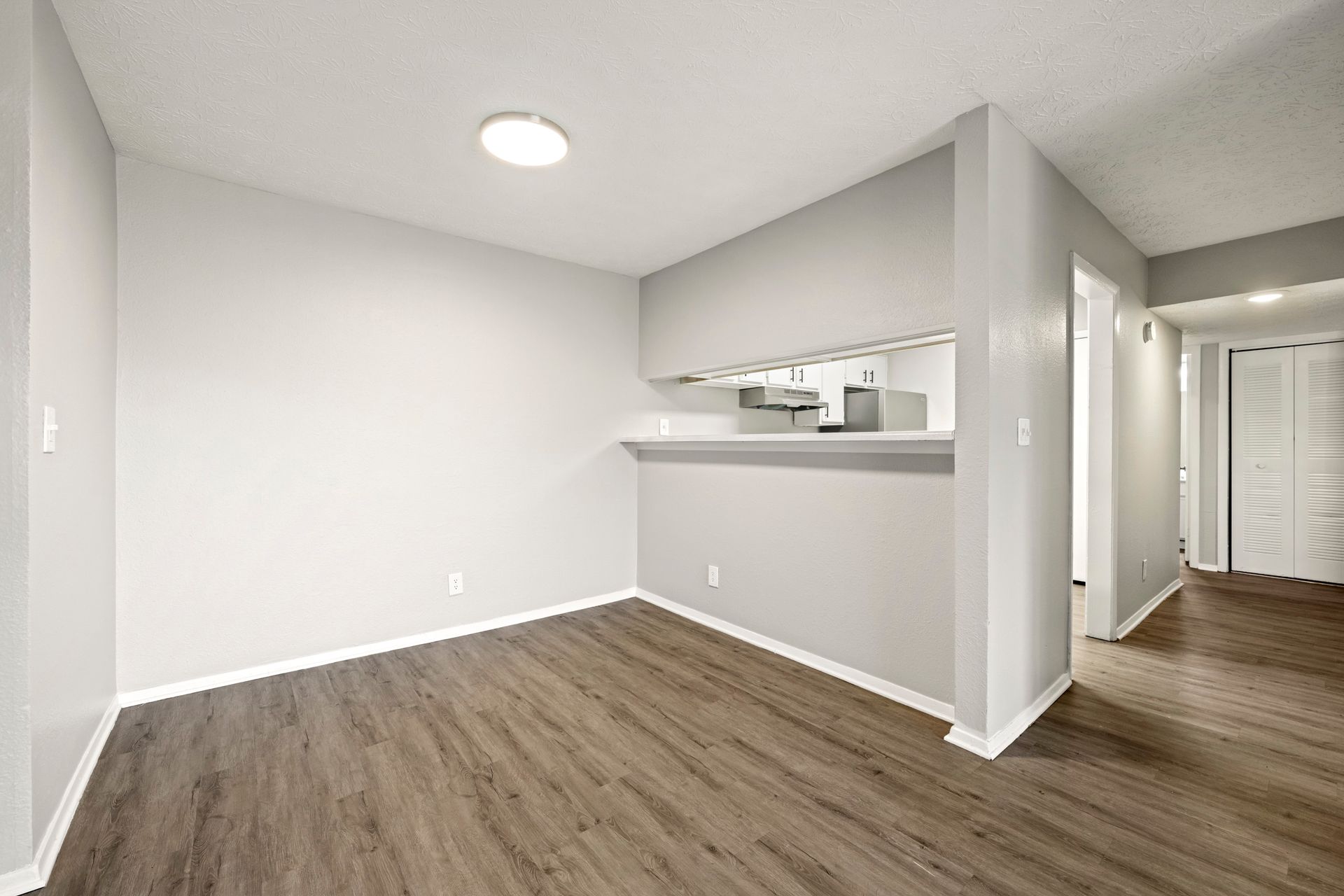 Empty modern apartment room with wood floors, white walls, and a pass-through to the kitchen.