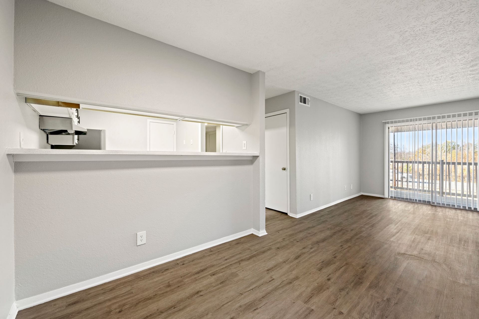 Empty apartment living room with wood floors, white walls, pass-through kitchen counter, and balcony doors.