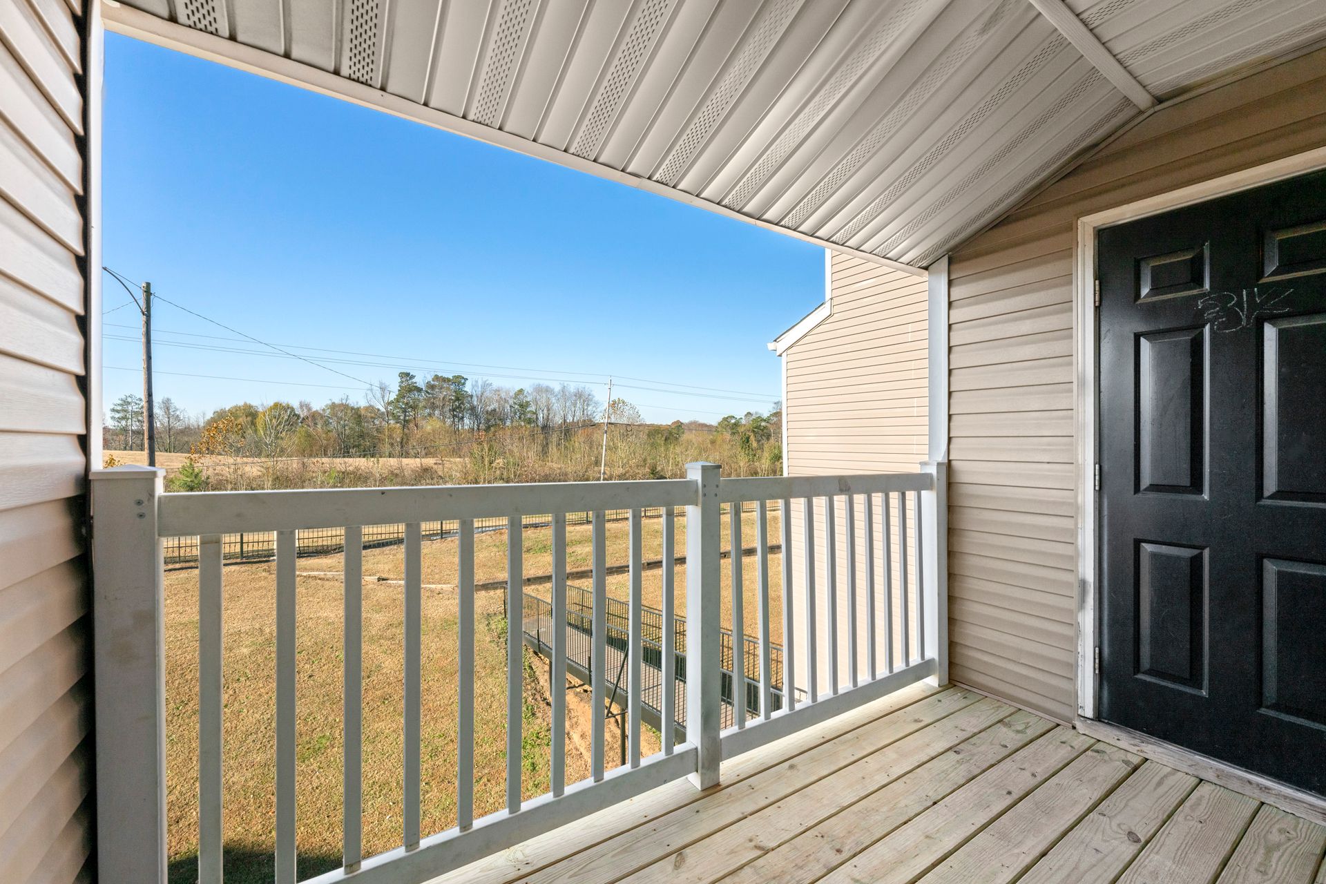 Covered front porch with white railing, black door, and view of open field under blue sky