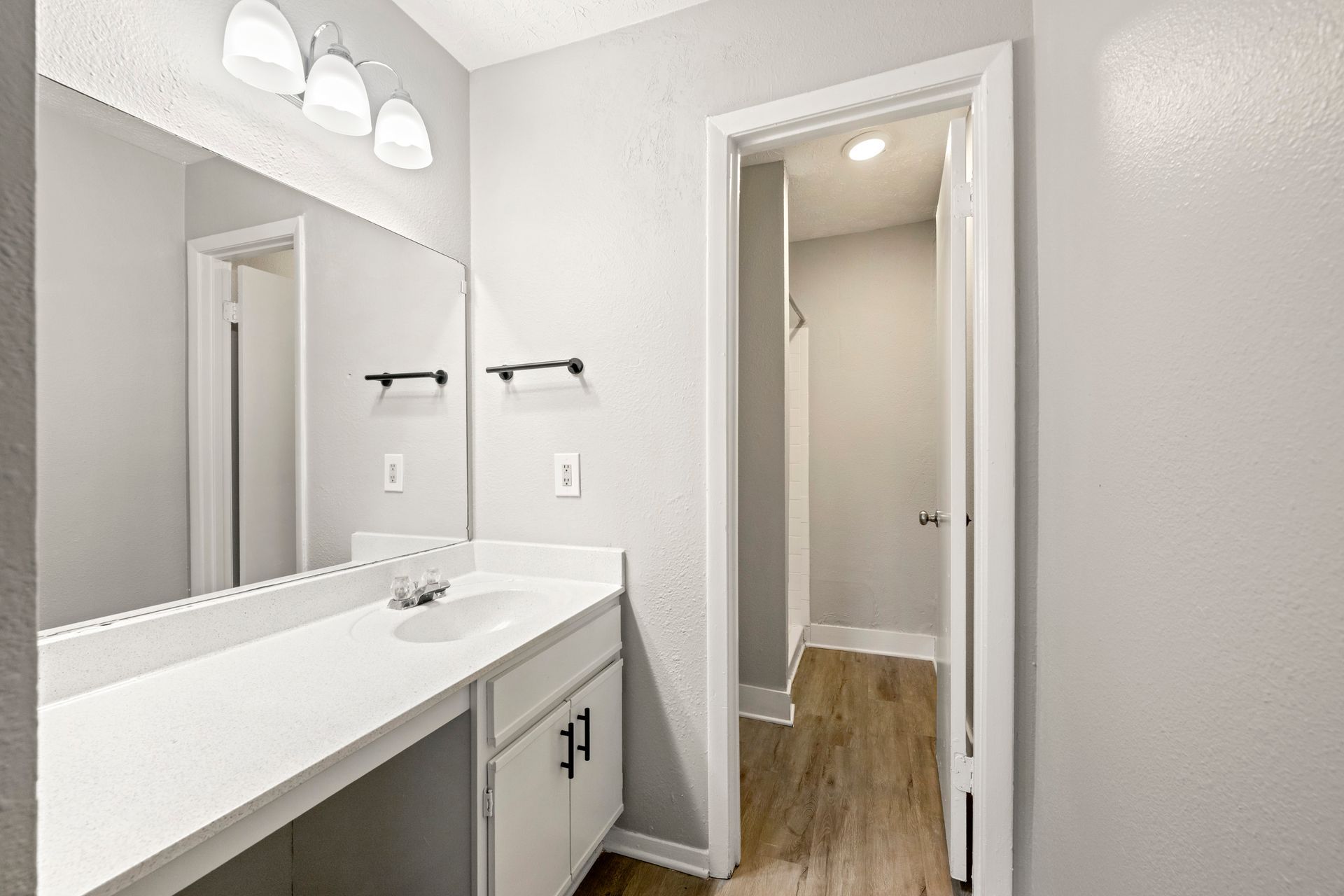 Bright bathroom vanity with large mirror, white cabinets, and a doorway to a wood-floored hall