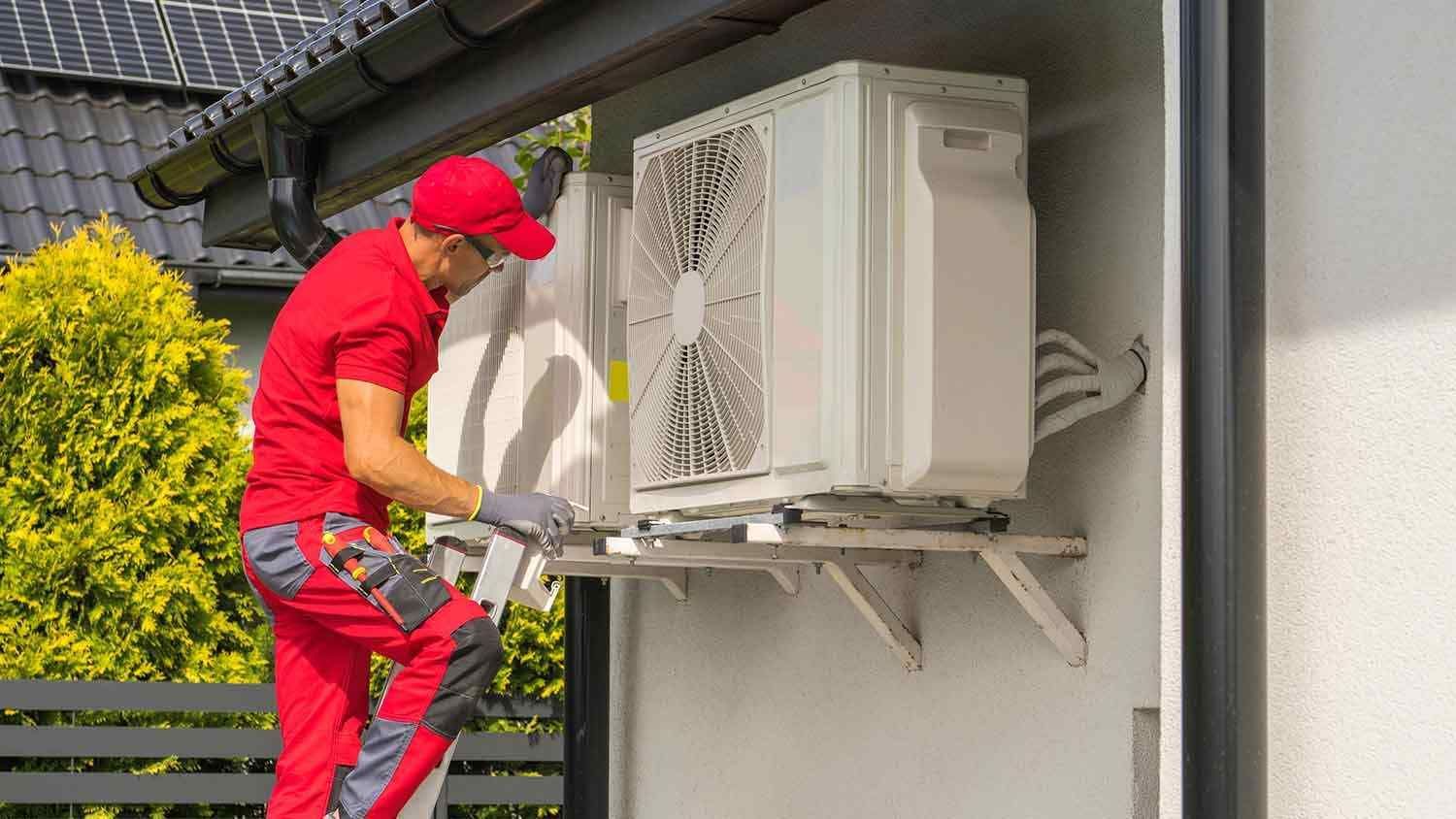 A worker in a red uniform stands on a ladder, installing an air conditioning unit on an exterior wall.