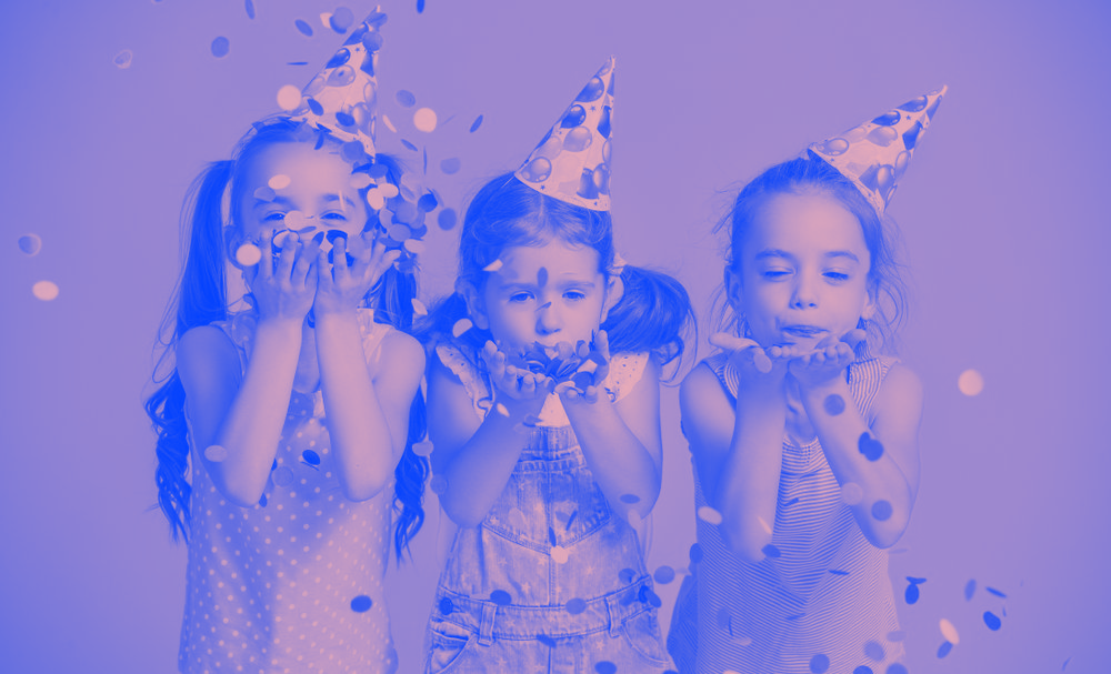 Three little girls wearing party hats are blowing confetti in the air.