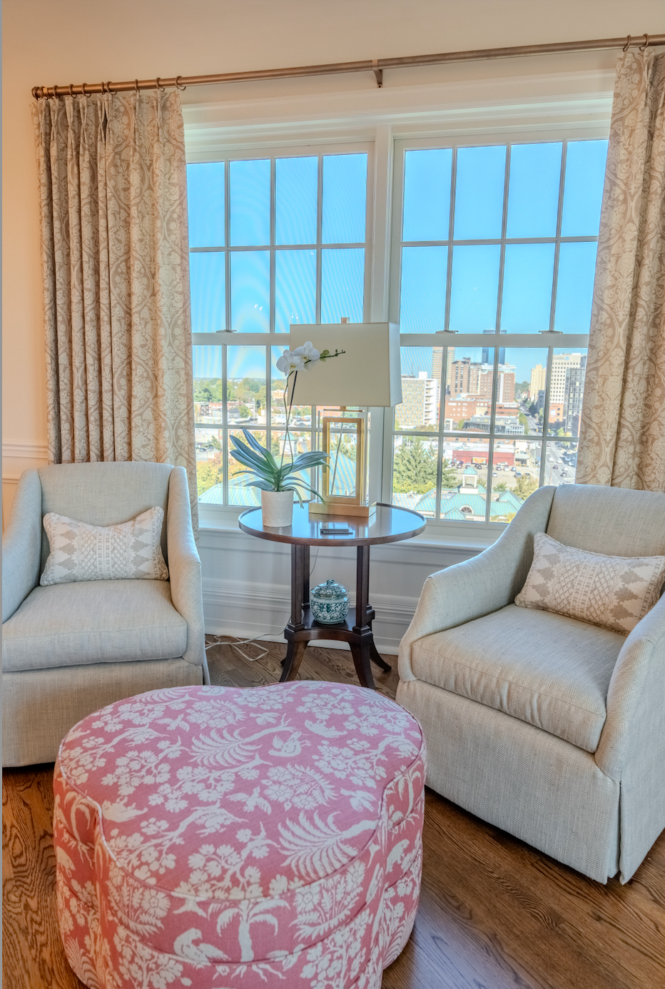 A living room with two chairs , an ottoman , a table and a window.