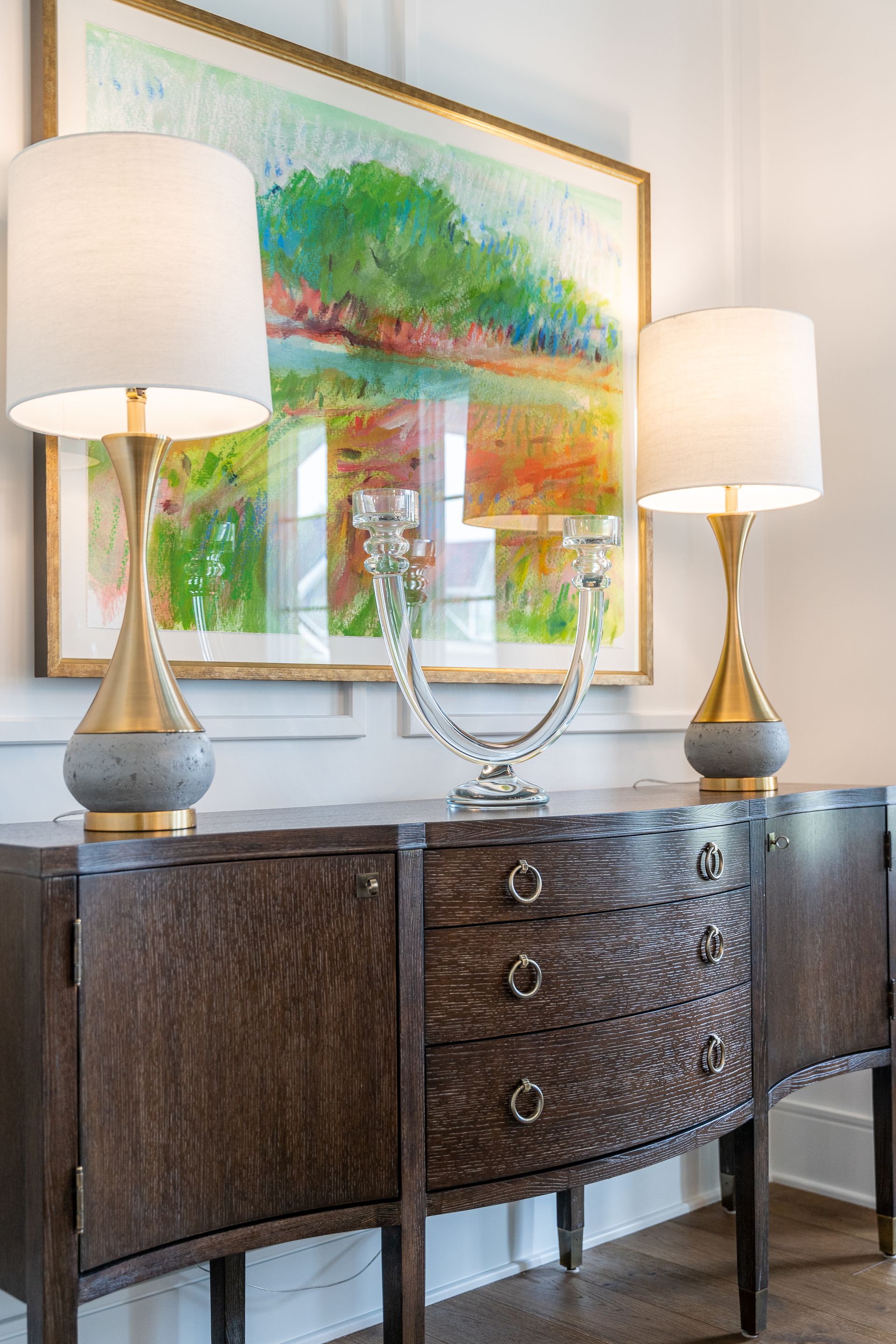 A wooden dresser with two lamps and a painting on the wall.