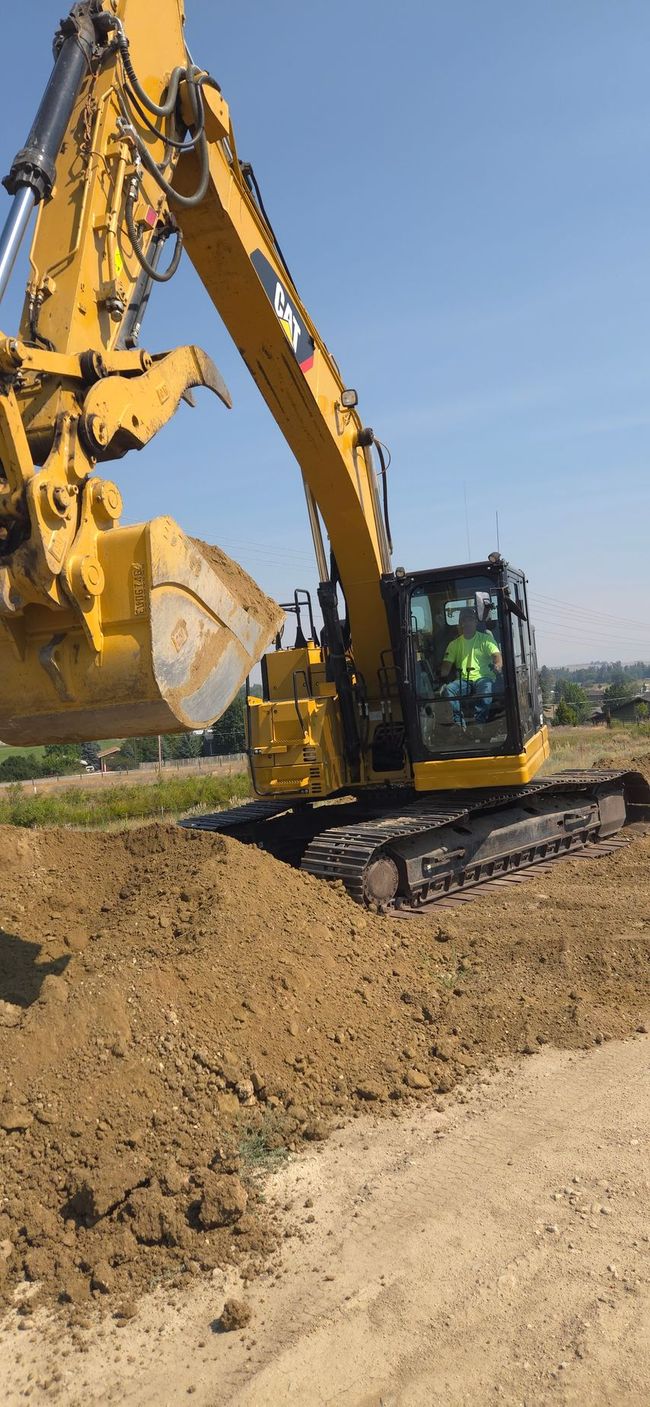 Excavator digging a dirt embankment while a worker stands nearby under a cloudy sky