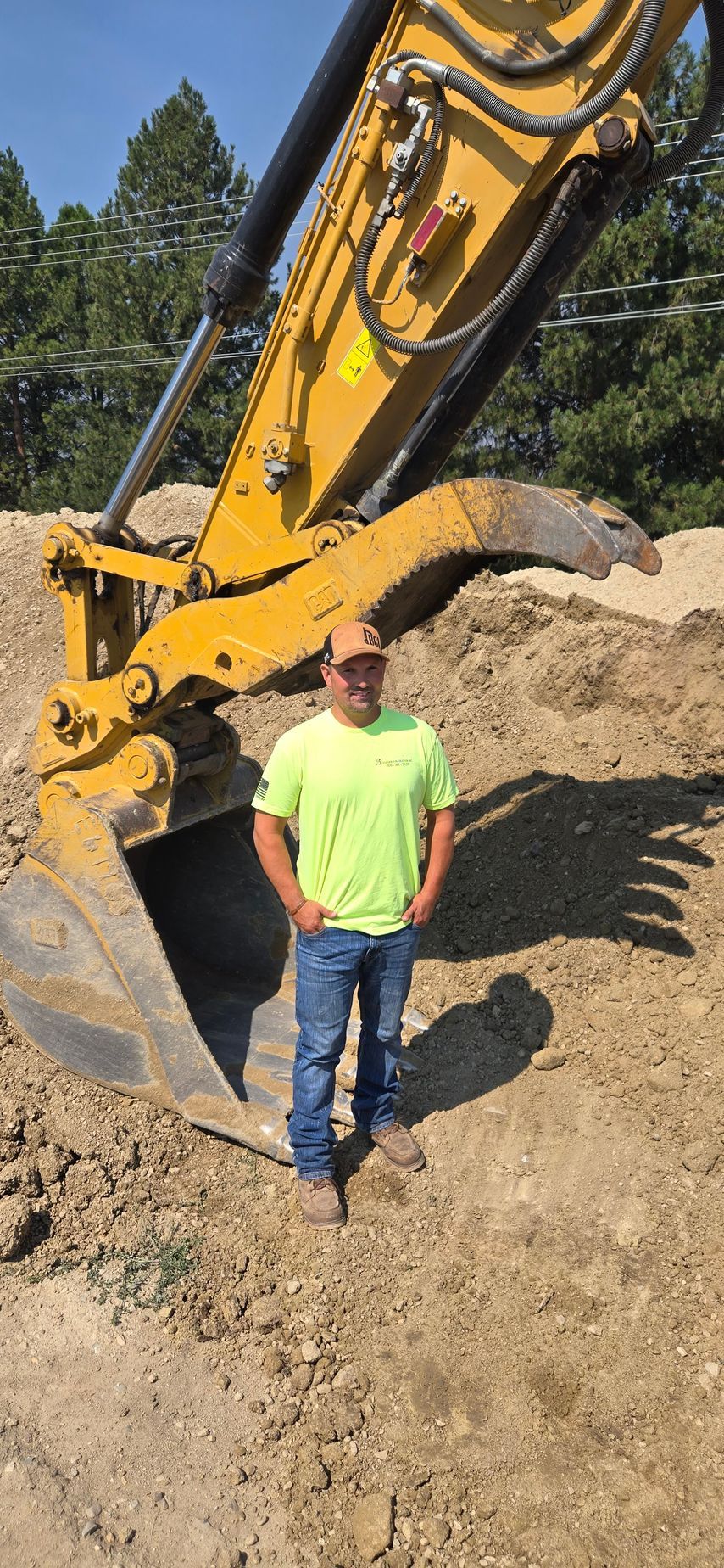 Man standing beside a yellow excavator bucket on a dirt construction site with sand piles and trees.