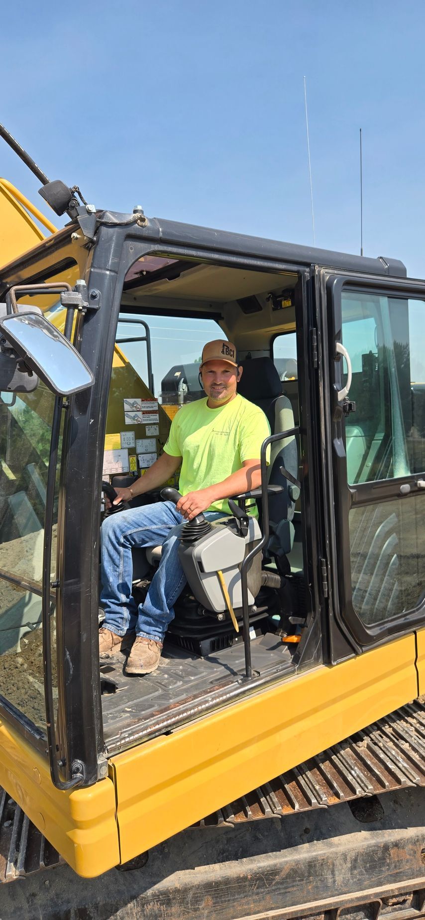 Two people seated in a yellow excavator cab, one in a neon green shirt, inside a construction site area