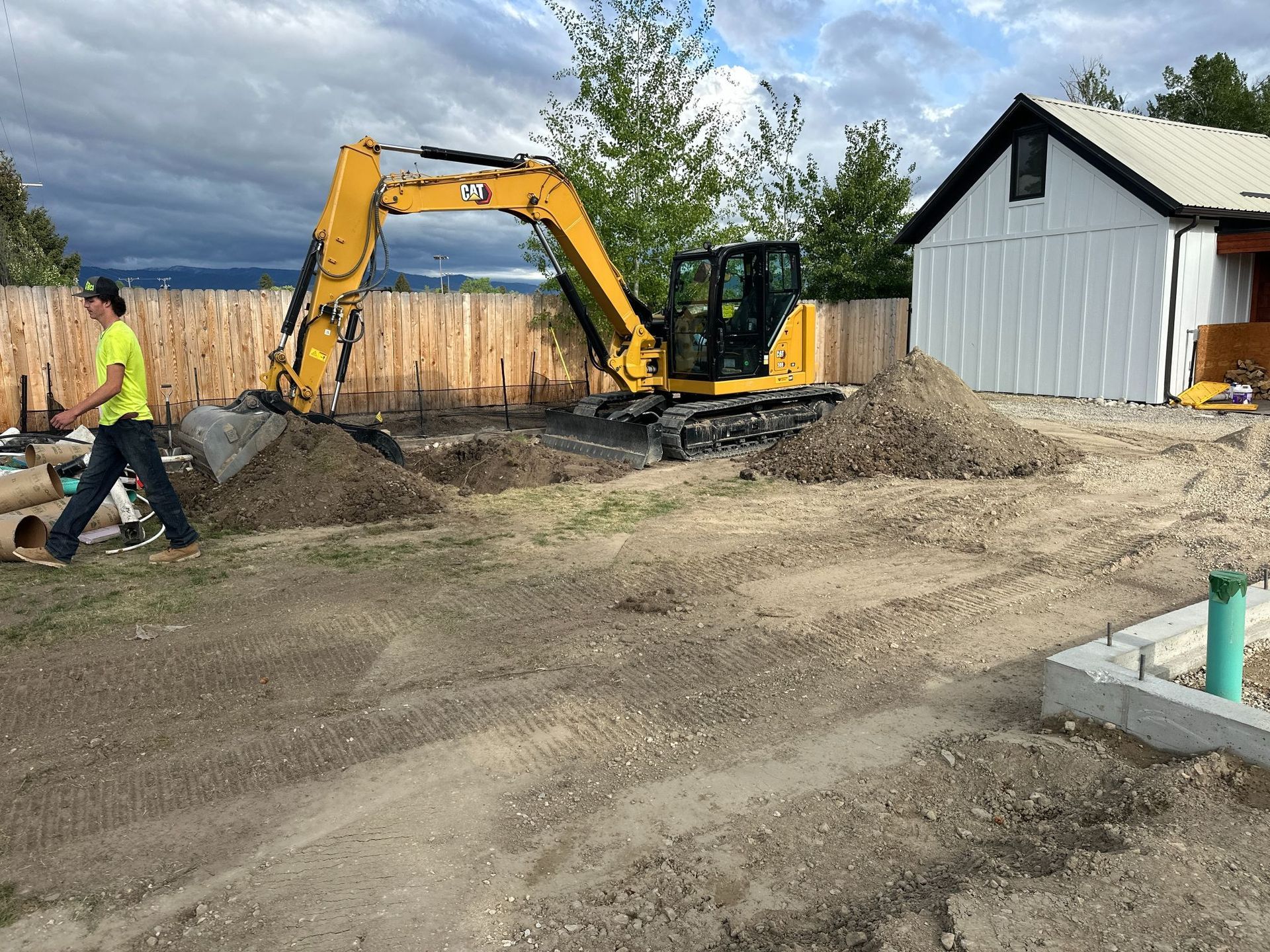 Excavator leveling dirt at a construction site beside a white building, with a worker walking nearby.