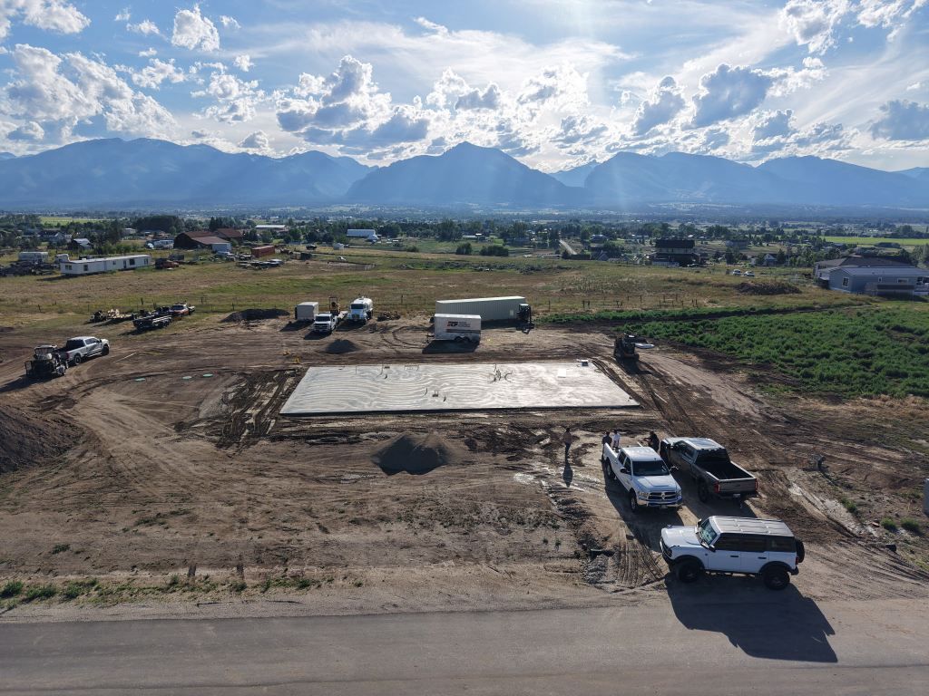 Construction site with concrete foundation, parked trucks, and mountains under a cloudy sky.