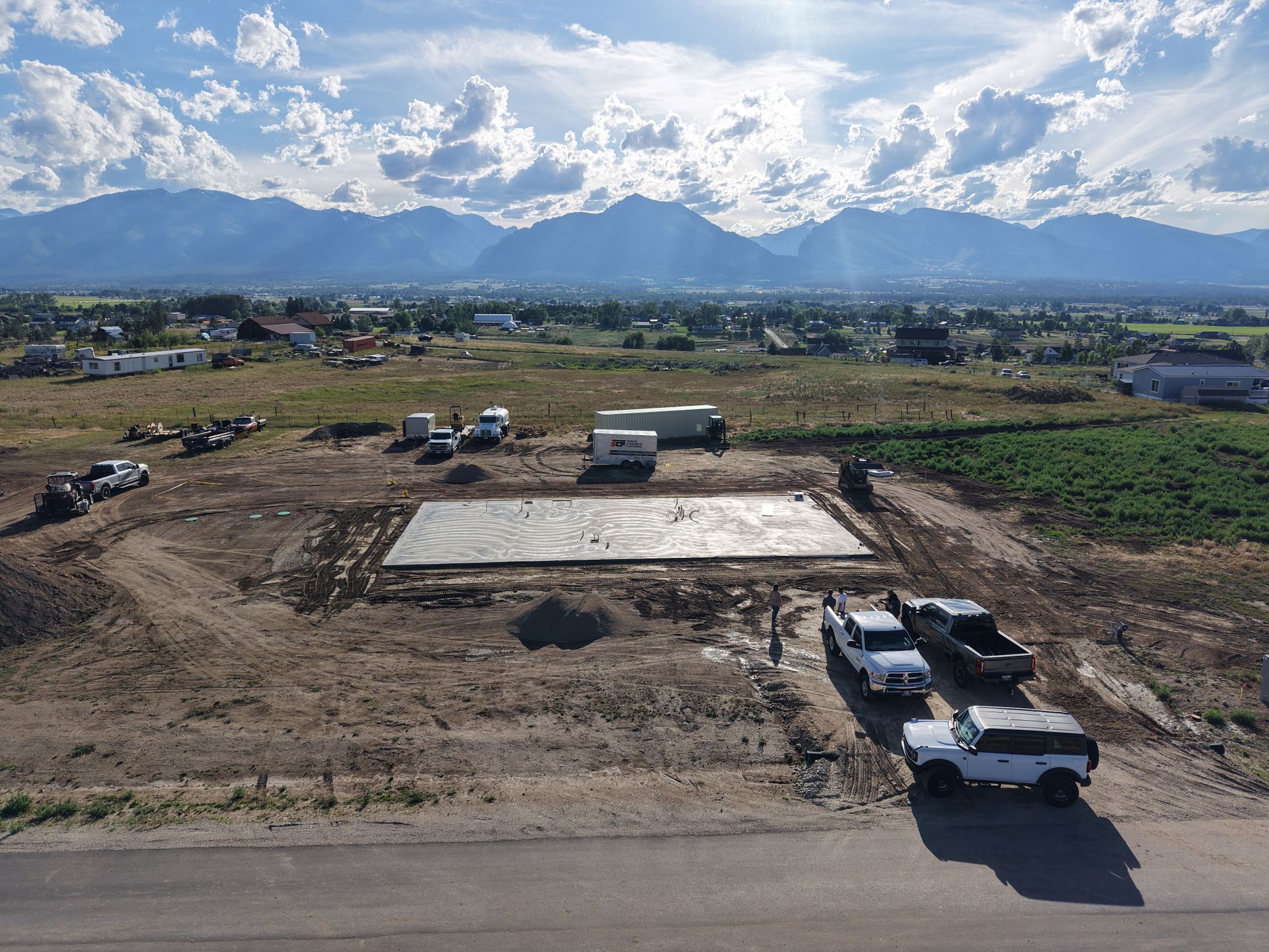 Rural construction site with a large concrete slab, trucks, and mountains under a cloudy sky