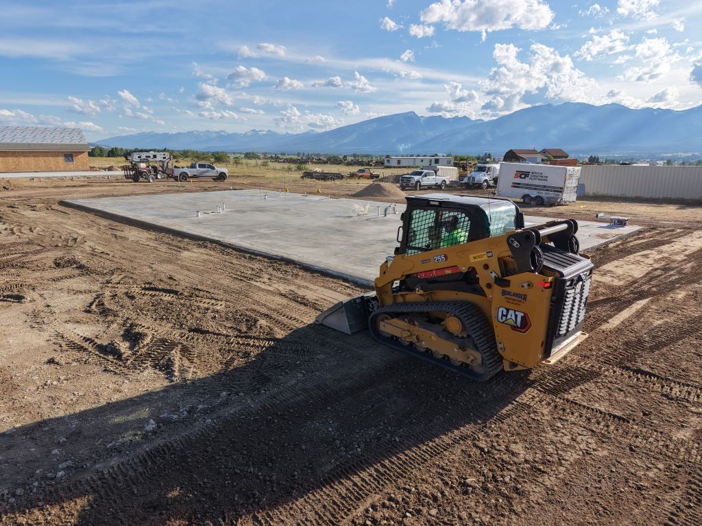 Yellow Caterpillar skid steer spreading black gravel on a construction site with mountains in the background