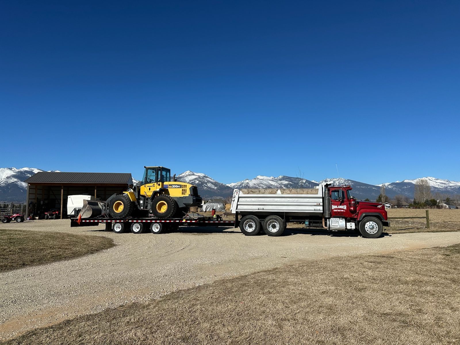 Red dump truck hauling a flatbed with a yellow loader in a gravel lot with mountains behind.