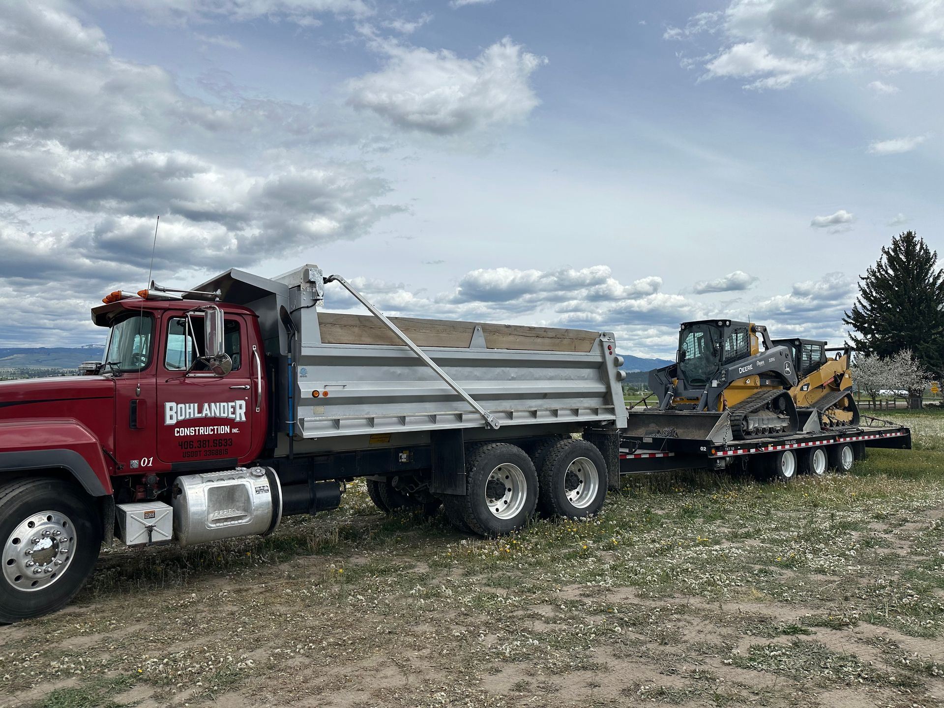Red tow truck hauling a flatbed trailer with a yellow excavator in a grassy field under a cloudy sky