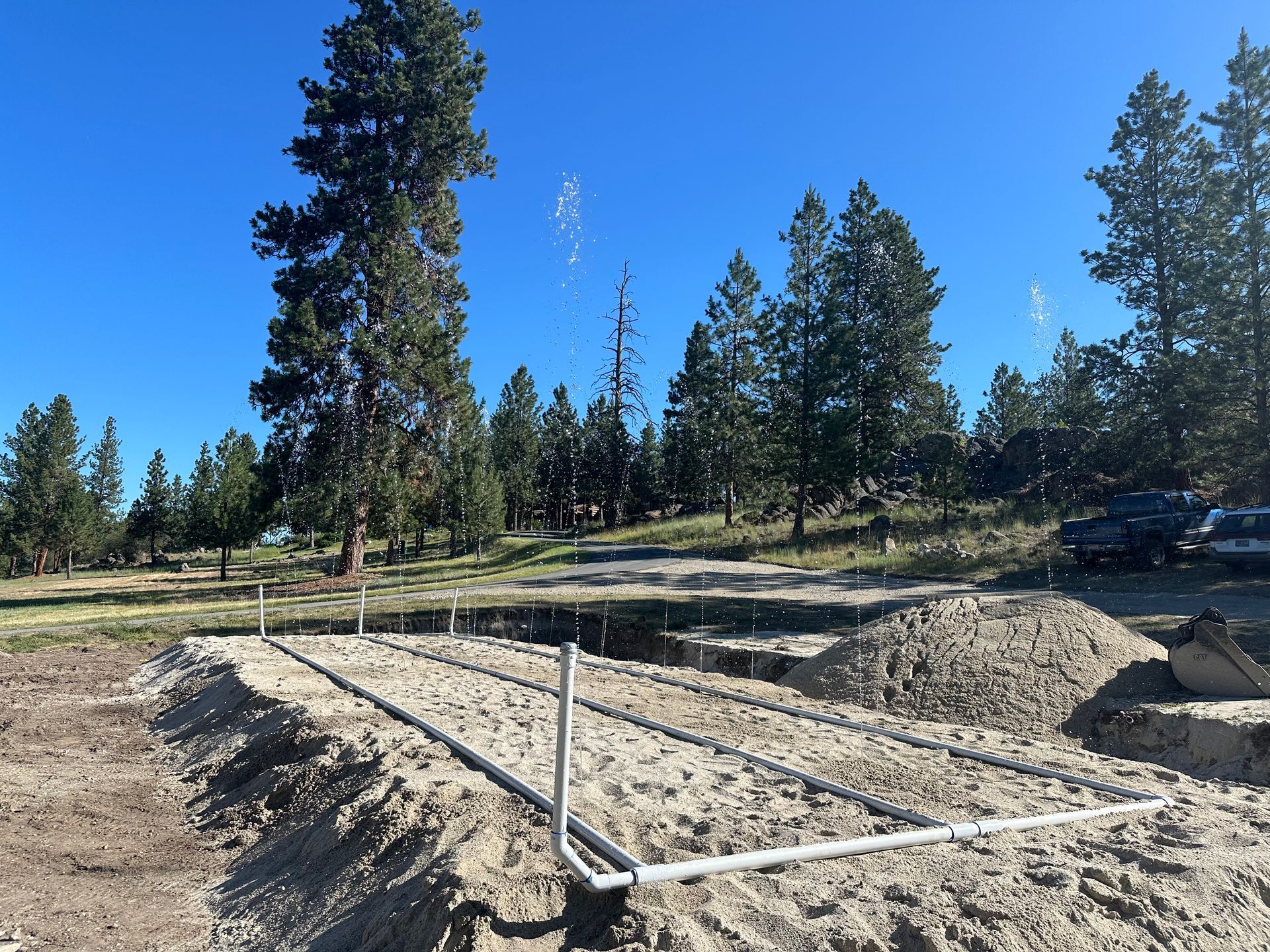 Cleared lot with white utility pipes and gravel under construction, surrounded by tall pine trees and blue sky