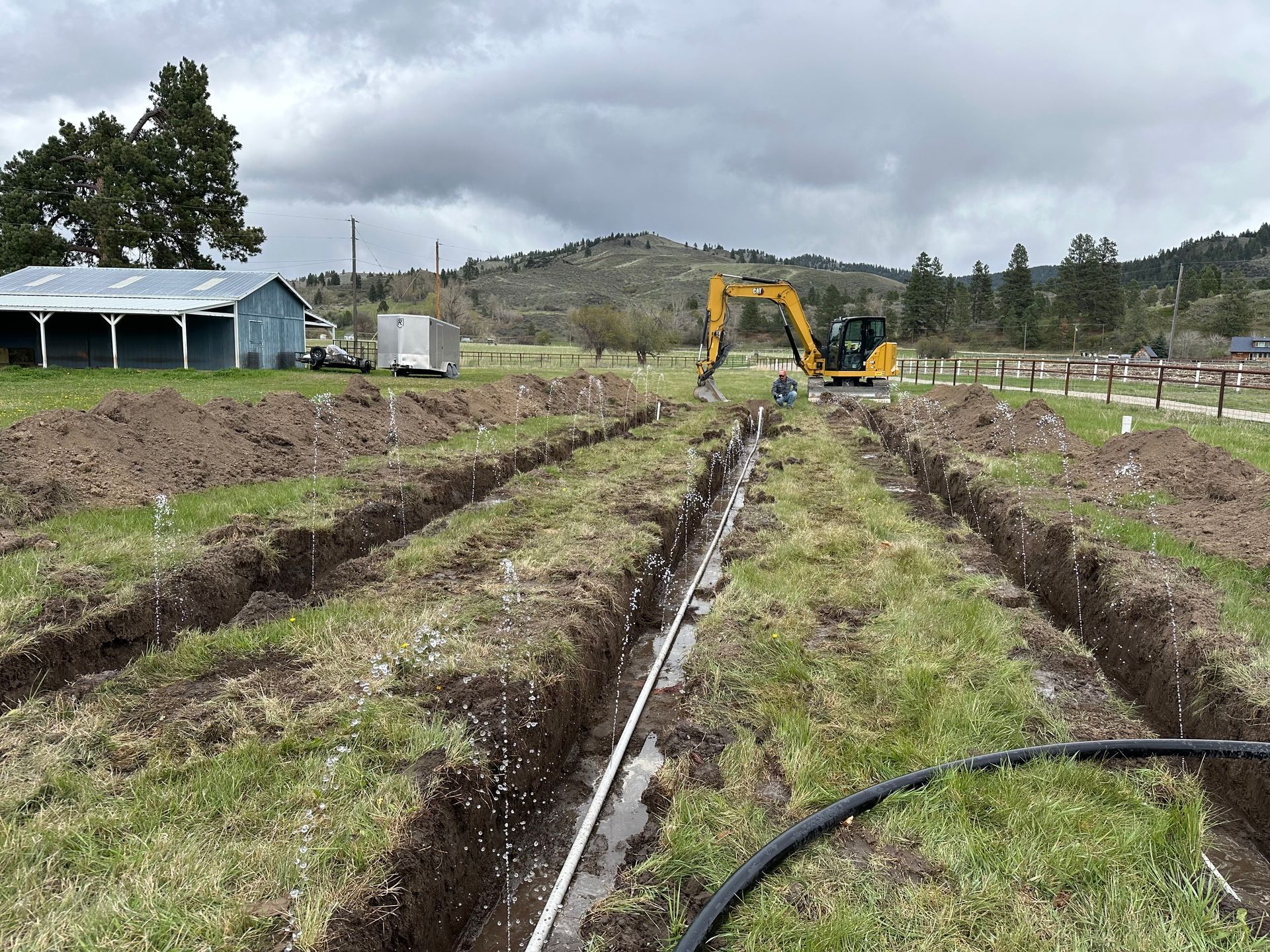 Excavator working in a muddy field with long trench rows and hoses under an overcast sky
