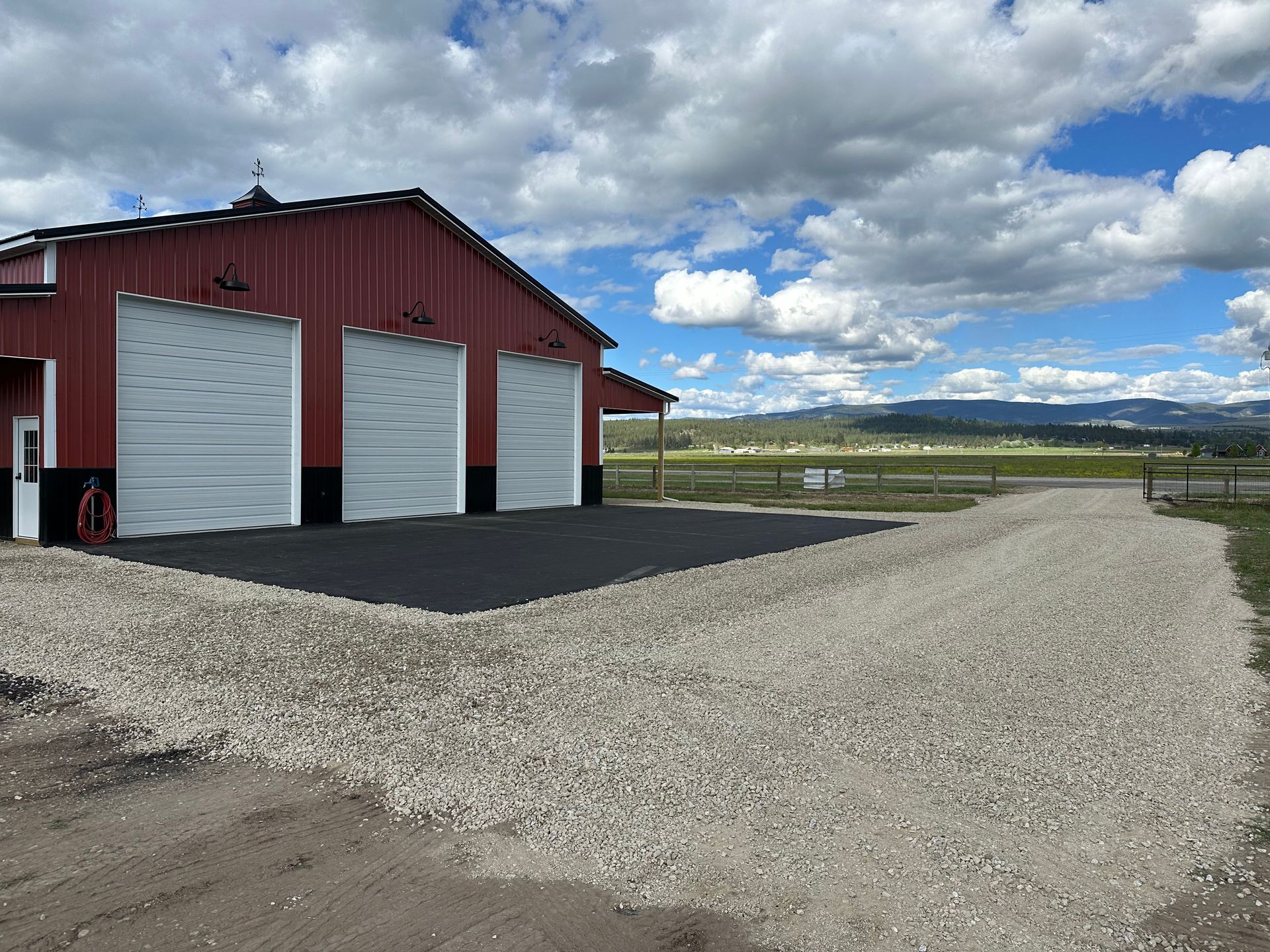 Red barn-style building with three white garage doors beside a gravel lot under a cloudy sky