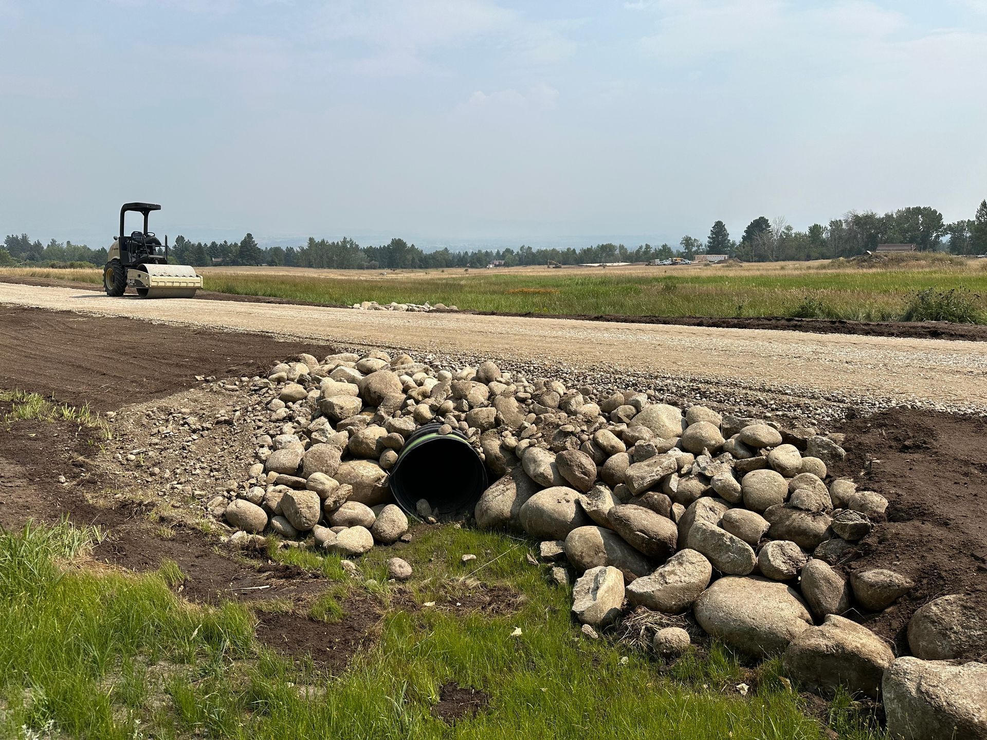 Construction site with a gravel road, rock-lined ditch, and small excavator in the distance