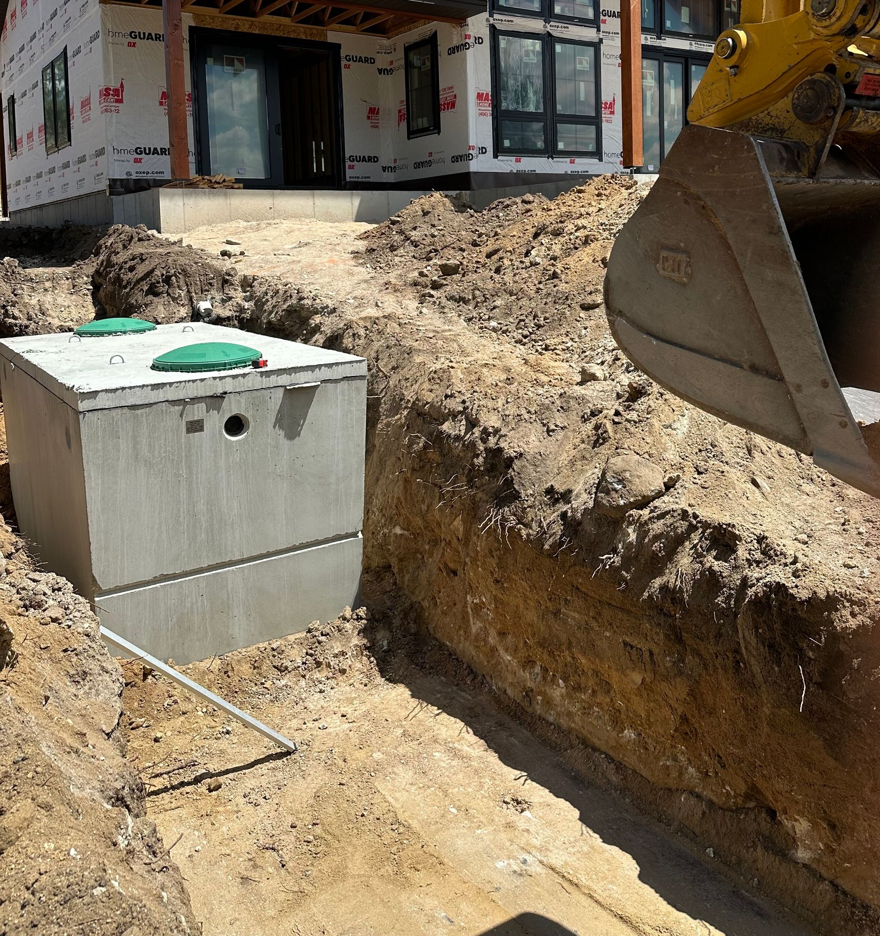 Construction trench with a concrete utility box and excavator bucket beside a building under construction
