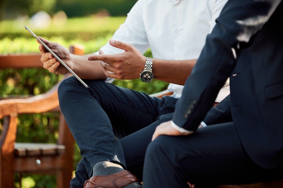 Two people in business attire look at a tablet together, seated on a wooden bench outside.