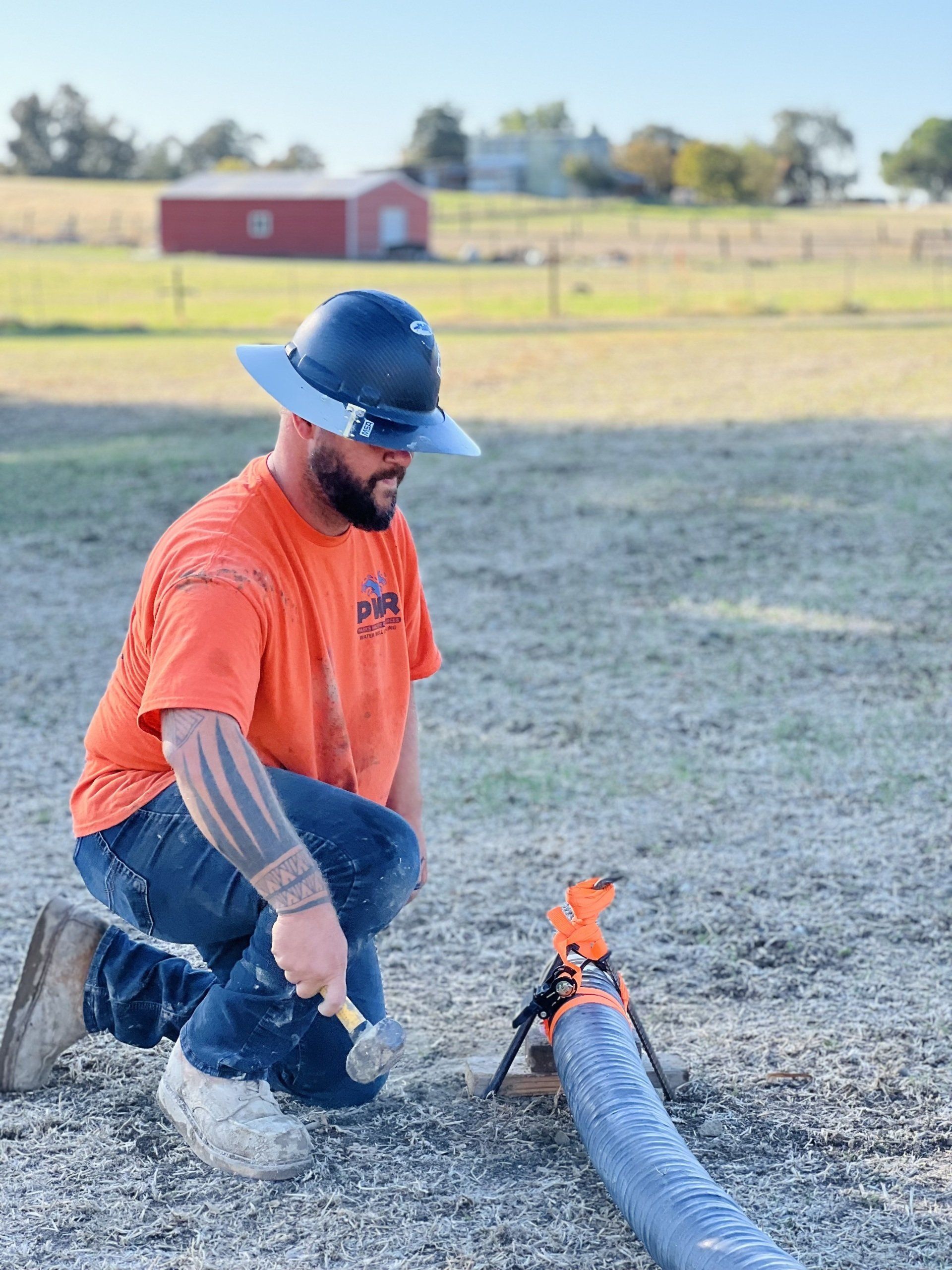 A man in an orange shirt and hard hat is kneeling down next to a hose.
