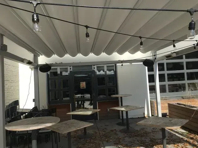 Outdoor patio with round tables under a white canopy, string lights, and a brick wall seating area