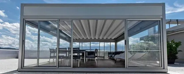Glass-walled modern patio room with open sliding doors and outdoor seating overlooking a bright view
