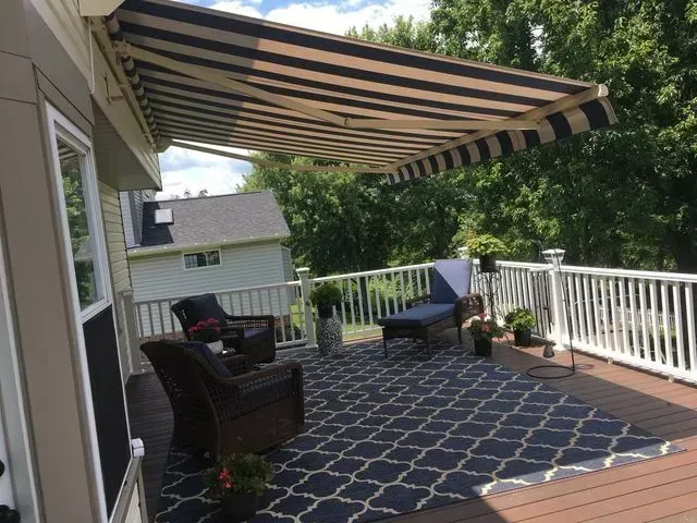 Covered backyard deck with striped awning, white railing, chairs, and patterned outdoor rug.