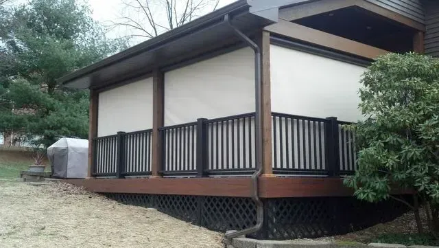 Small house with a covered porch, dark railings, and light exterior walls in a wooded yard.
