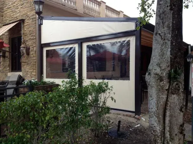 Small glass sunroom addition beside a brick house, with dark trim and shrubs in front.