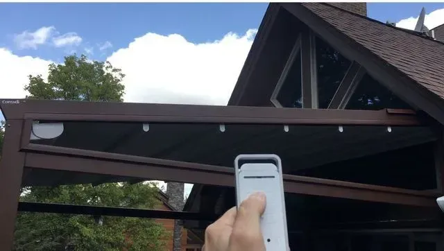 Hand holding a white remote pointing at a house roof and wooden pergola under a cloudy sky