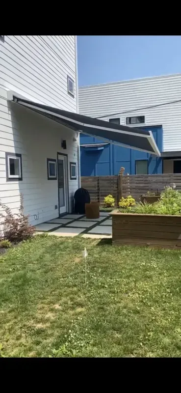 Side entrance of a white house with awning, blue door, small garden bed, and lawn under a clear sky