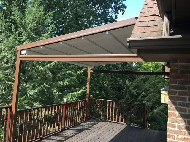 Covered wooden deck with railing and angled translucent awning beside a brick house, surrounded by trees
