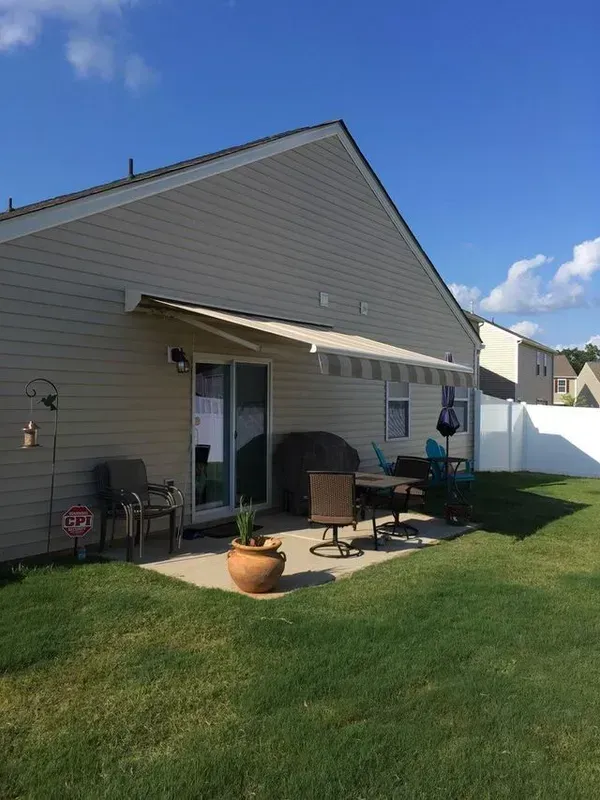 Back patio with chairs, grill, potted plant, and patio cover beside a grassy yard under a blue sky