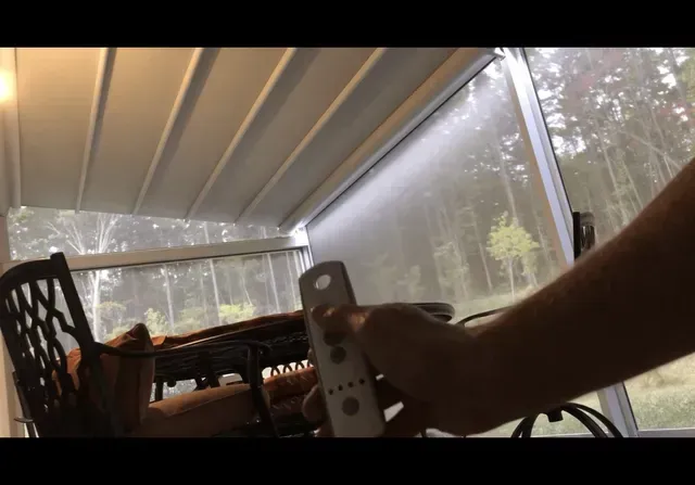 Person holding a remote on a screened porch with chairs and a wooded view.