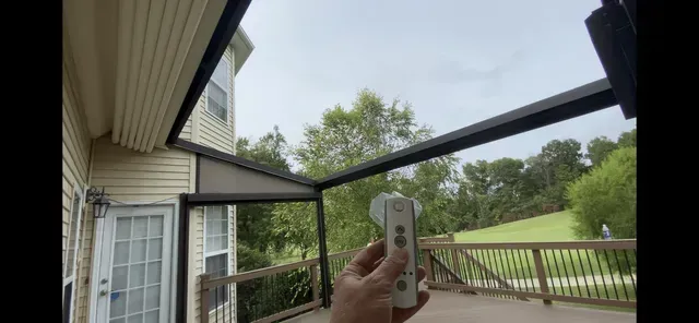 Hand holding a remote on a screened porch overlooking a green backyard and trees