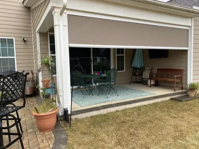 Covered patio with outdoor table, chairs, and bench beside a beige house and lawn