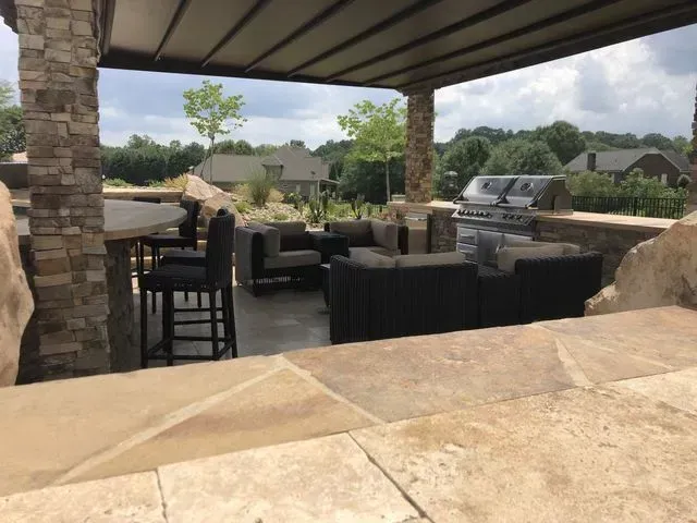 Covered backyard patio with stone seating, outdoor furniture, grill, and landscaped neighborhood view