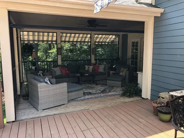 Covered patio with wicker seating, rug, and potted plants beside a wooden deck.